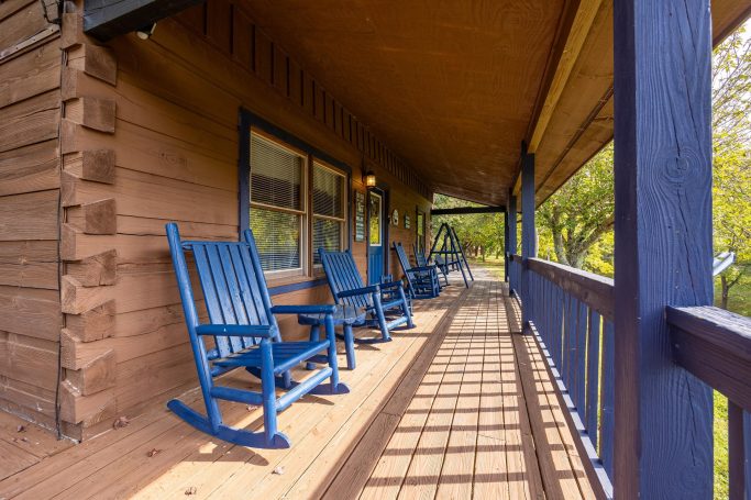 Blue rocking chairs line a wooden porch with sunlight casting shadows.