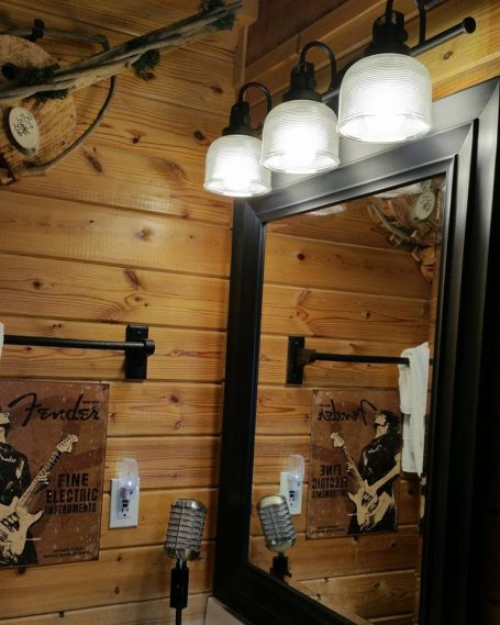 Bathroom mirror with rustic wooden walls, three light fixtures, and decorative items.