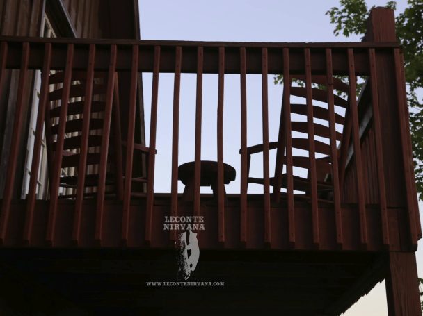 Two rocking chairs and a small table on a wooden balcony with trees in the background.